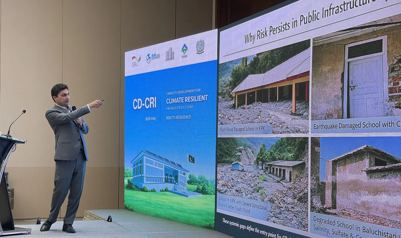 A speaker presenting CD-CRI slides on why risk persists in public infrastructure systems, beside the programme's title slide.