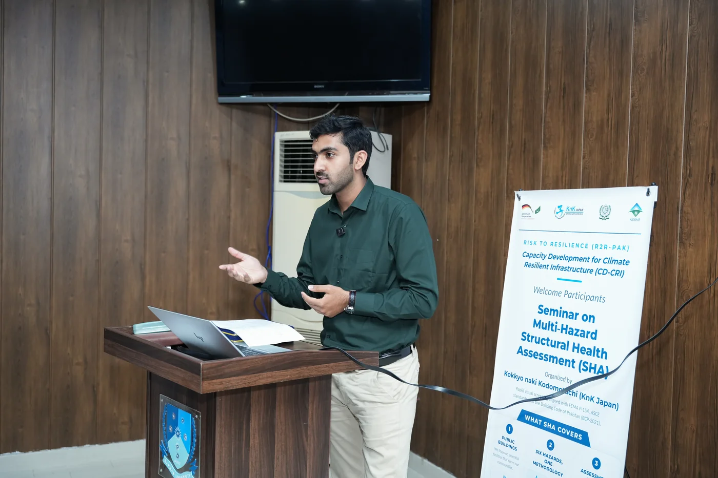 A presenter introduces the Multi-Hazard Structural Health Assessment method beside a CD-CRI seminar banner at a university campus.