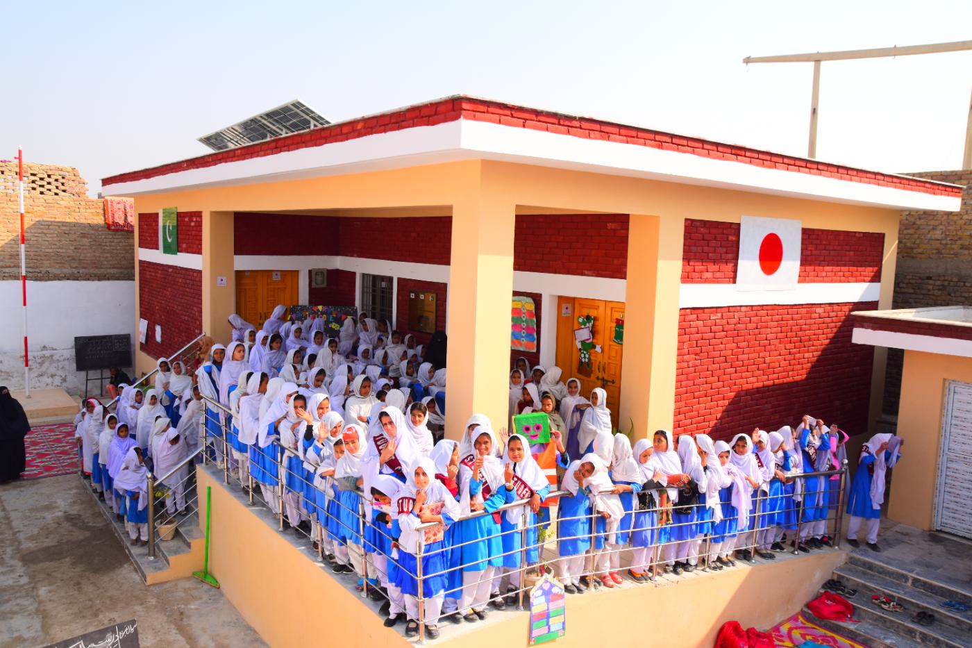 Girls in white and blue uniforms gathered at a newly built KnK Japan school with Pakistan and Japan flags