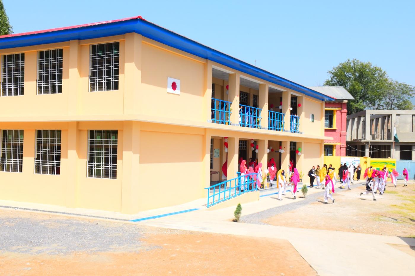 Two-storey KnK Japan school GGHS Oghi with blue roofing, accessibility ramp and students at handover ceremony
