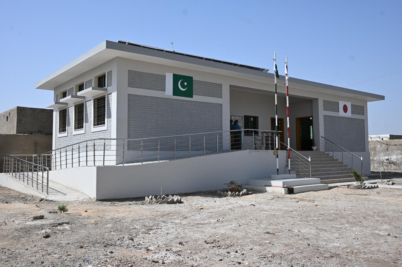 Newly built grey-brick Basic Health Unit with Pakistan and Japan flags, an accessibility ramp and rooftop solar