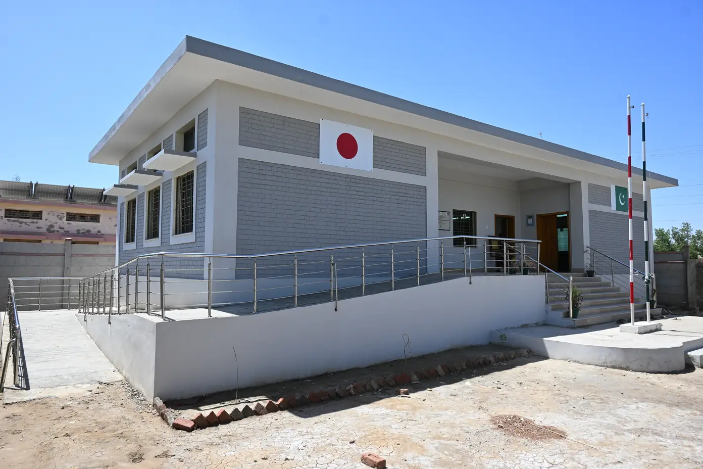 Grey-brick KnK Japan Basic Health Unit with a full-length wheelchair ramp, Japan and Pakistan flags