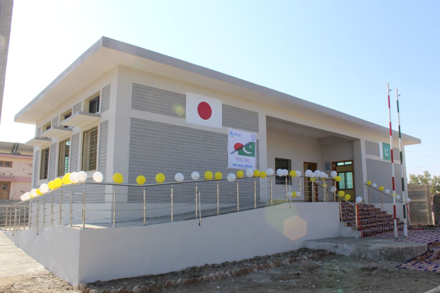 Newly reconstructed grey-brick Basic Health Unit with Japan flag, accessibility ramp and balloons at handover ceremony