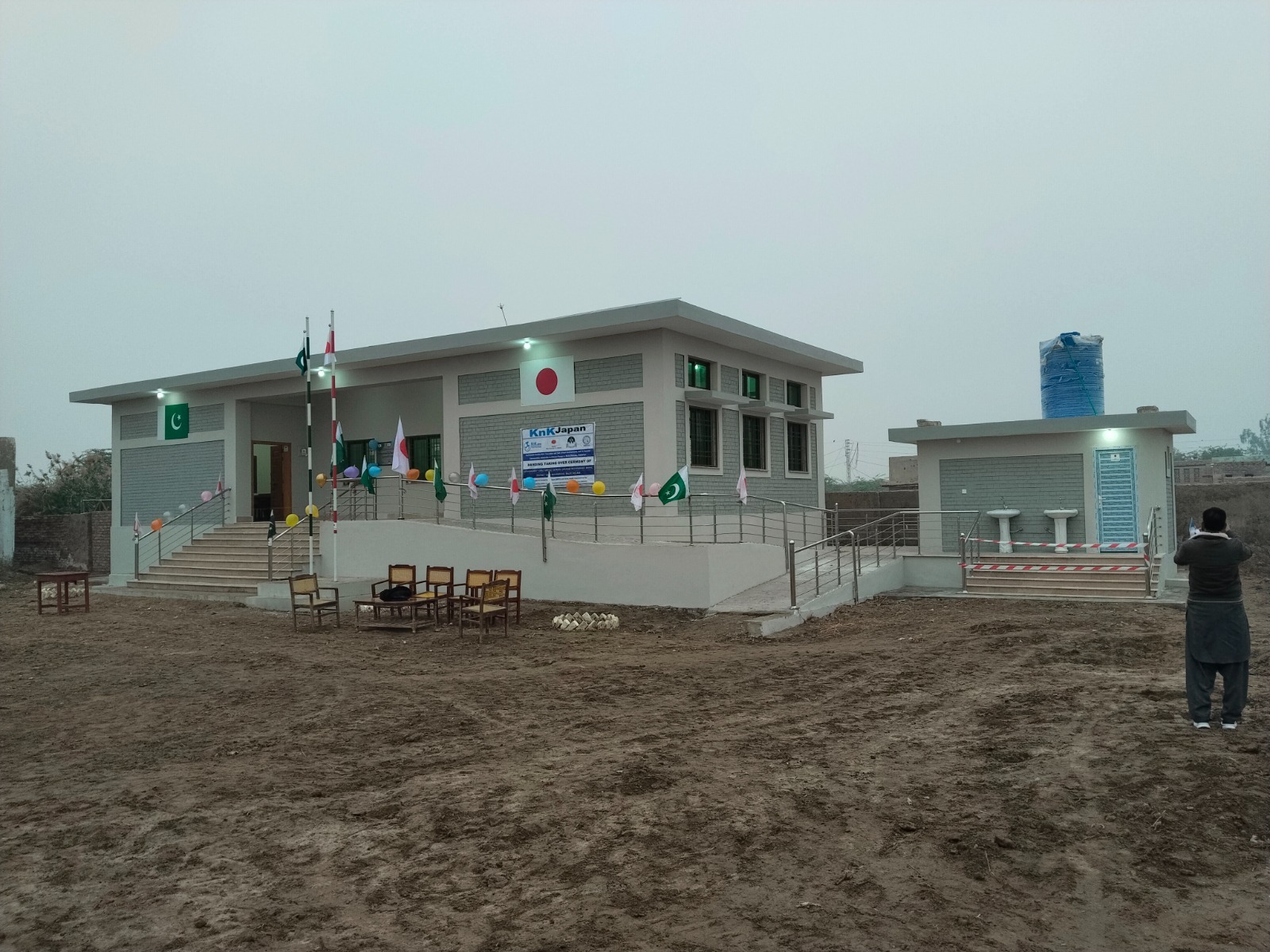Handover ceremony at a newly built KnK Japan Basic Health Unit at dusk, with chairs, banner and flags