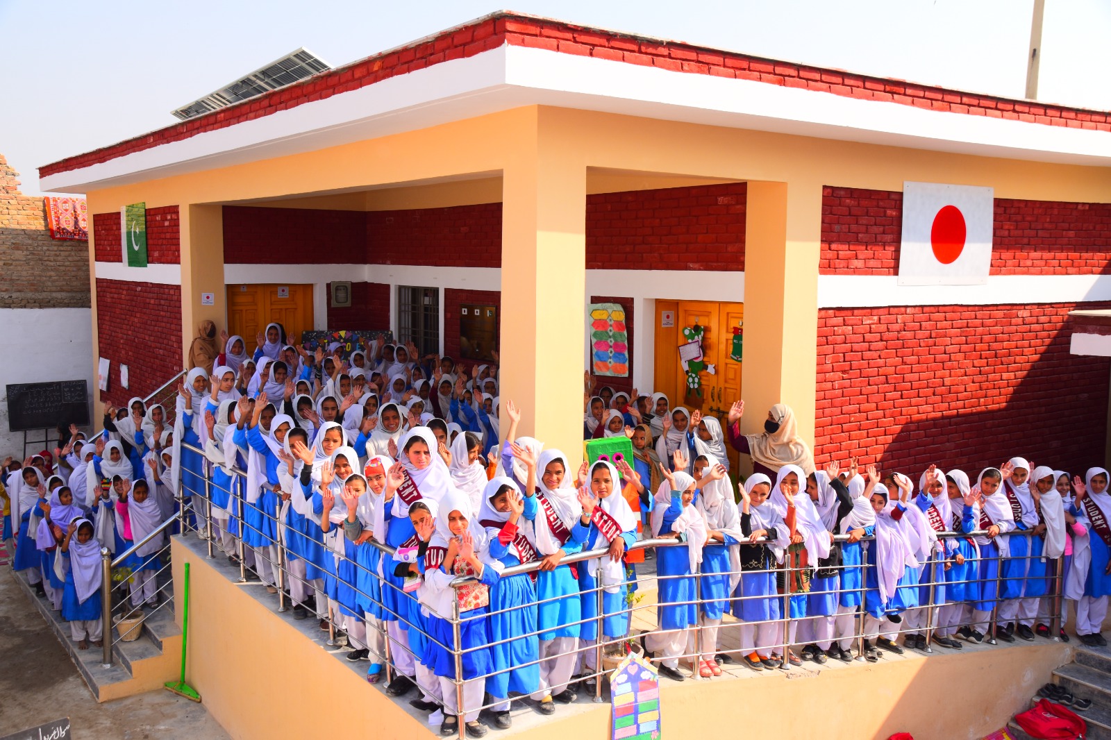 Students waving from the upper deck of a KnK Japan school in Pakistan, with the Pakistan and Japan flags painted on the wall